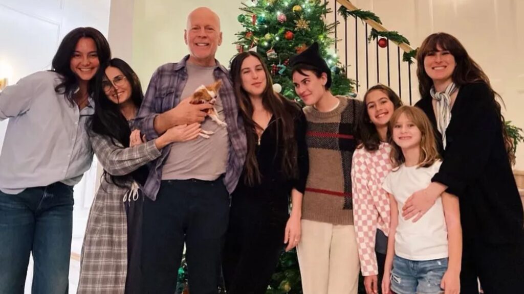 A imagem mostra Bruce Willis ao lado de sua esposa Emma Heming, de sua ex-esposa Demi Moore e de suas cinco filhas, reunidos em frente a uma árvore de Natal decorada. Todos estão abraçados e sorridentes, transmitindo um clima de união e alegria familiar. Bruce segura um pequeno cachorro nos braços, enquanto os demais posam próximos, reforçando o espírito de afeto e proximidade. 