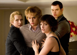 A imagem mostra quatro personagens da saga Crepúsculo, em um momento tenso e de aparente alerta. Eles estão próximos uns dos outros, com expressões sérias e protetoras. À esquerda, um homem loiro de expressão preocupada segura outro rapaz loiro no centro, que parece assustado. À frente, uma jovem de cabelos curtos e escuros olha atentamente para fora da cena, enquanto outro homem forte e de olhar fixo se posiciona atrás dela, também em postura protetora. O ambiente é interno, iluminado, com um arranjo de flores ao fundo. A cena transmite uma sensação de urgência e união entre os personagens