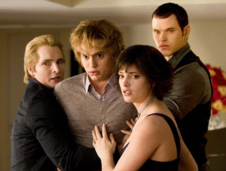 A imagem mostra quatro personagens da saga Crepúsculo, em um momento tenso e de aparente alerta. Eles estão próximos uns dos outros, com expressões sérias e protetoras. À esquerda, um homem loiro de expressão preocupada segura outro rapaz loiro no centro, que parece assustado. À frente, uma jovem de cabelos curtos e escuros olha atentamente para fora da cena, enquanto outro homem forte e de olhar fixo se posiciona atrás dela, também em postura protetora. O ambiente é interno, iluminado, com um arranjo de flores ao fundo. A cena transmite uma sensação de urgência e união entre os personagens
