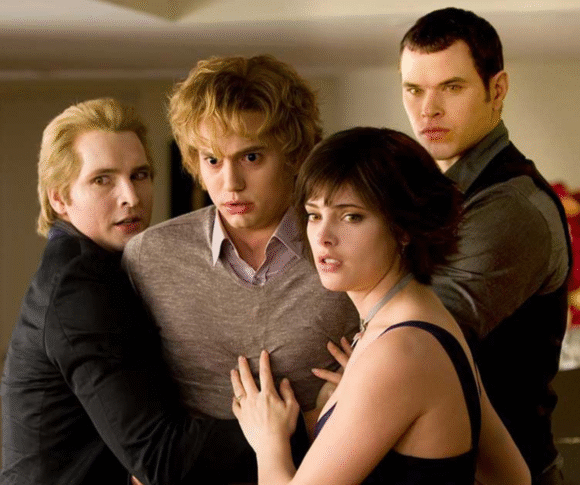 A imagem mostra quatro personagens da saga Crepúsculo, em um momento tenso e de aparente alerta. Eles estão próximos uns dos outros, com expressões sérias e protetoras. À esquerda, um homem loiro de expressão preocupada segura outro rapaz loiro no centro, que parece assustado. À frente, uma jovem de cabelos curtos e escuros olha atentamente para fora da cena, enquanto outro homem forte e de olhar fixo se posiciona atrás dela, também em postura protetora. O ambiente é interno, iluminado, com um arranjo de flores ao fundo. A cena transmite uma sensação de urgência e união entre os personagens