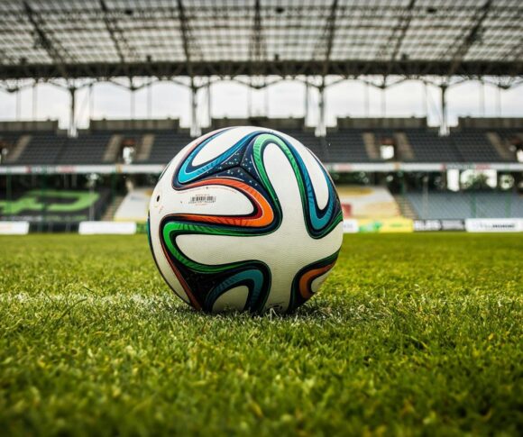 A imagem mostra uma bola de futebol em destaque, posicionada sobre a grama verde de um campo. Ao fundo, aparece parte de um estádio com arquibancadas vazias e uma cobertura metálica. O céu está nublado, criando uma iluminação suave sobre a cena. A bola tem detalhes coloridos em azul, verde, laranja e preto, contrastando com o branco de sua superfície