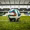 A imagem mostra uma bola de futebol em destaque, posicionada sobre a grama verde de um campo. Ao fundo, aparece parte de um estádio com arquibancadas vazias e uma cobertura metálica. O céu está nublado, criando uma iluminação suave sobre a cena. A bola tem detalhes coloridos em azul, verde, laranja e preto, contrastando com o branco de sua superfície