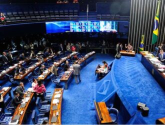 A imagem mostra o plenário do Senado Federal, em Brasília, com carpete azul e mesas de madeira em formato semicircular. Senadores e assessores conversam e trabalham, enquanto a mesa diretora aparece ao fundo, diante da bandeira do Brasil. Painéis eletrônicos exibem informações da sessão, e no nível superior há lugares para público e imprensa, compondo um cenário formal e movimentado