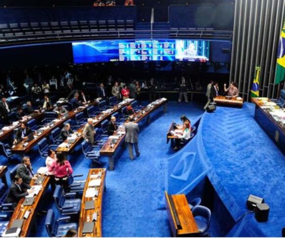 A imagem mostra o plenário do Senado Federal, em Brasília, com carpete azul e mesas de madeira em formato semicircular. Senadores e assessores conversam e trabalham, enquanto a mesa diretora aparece ao fundo, diante da bandeira do Brasil. Painéis eletrônicos exibem informações da sessão, e no nível superior há lugares para público e imprensa, compondo um cenário formal e movimentado