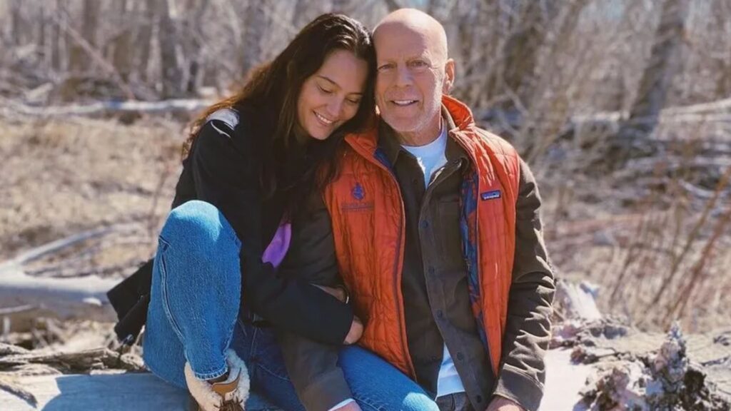 A foto mostra Emma Heming e Bruce Willis sentados lado a lado em um ambiente ao ar livre, possivelmente em meio a árvores durante o inverno ou início da primavera. Emma está encostada carinhosamente no ombro de Bruce, sorrindo de olhos fechados, enquanto ele olha para a câmera com uma expressão serena. Bruce veste uma camisa escura com colete laranja, e Emma usa um casaco preto e calça jeans. A cena transmite proximidade, afeto e tranquilidade.