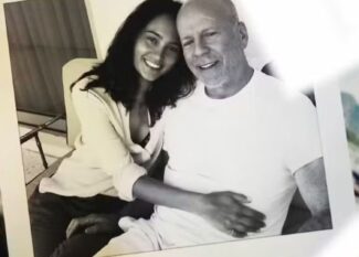 A foto em preto e branco mostra Emma Heming e Bruce Willis sentados lado a lado, sorridentes. Emma está encostada em Bruce, abraçando-o pela cintura em um gesto de carinho e proximidade. Ambos transmitem afeto e serenidade. A imagem aparece impressa, posicionada sobre uma superfície junto a outras fotografias parcialmente visíveis ao fundo