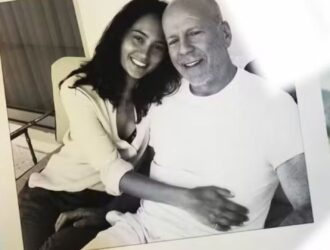 A foto em preto e branco mostra Emma Heming e Bruce Willis sentados lado a lado, sorridentes. Emma está encostada em Bruce, abraçando-o pela cintura em um gesto de carinho e proximidade. Ambos transmitem afeto e serenidade. A imagem aparece impressa, posicionada sobre uma superfície junto a outras fotografias parcialmente visíveis ao fundo