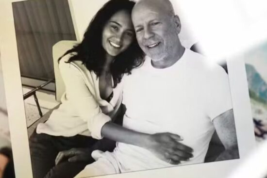 A foto em preto e branco mostra Emma Heming e Bruce Willis sentados lado a lado, sorridentes. Emma está encostada em Bruce, abraçando-o pela cintura em um gesto de carinho e proximidade. Ambos transmitem afeto e serenidade. A imagem aparece impressa, posicionada sobre uma superfície junto a outras fotografias parcialmente visíveis ao fundo