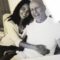 A foto em preto e branco mostra Emma Heming e Bruce Willis sentados lado a lado, sorridentes. Emma está encostada em Bruce, abraçando-o pela cintura em um gesto de carinho e proximidade. Ambos transmitem afeto e serenidade. A imagem aparece impressa, posicionada sobre uma superfície junto a outras fotografias parcialmente visíveis ao fundo