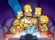A imagem mostra a família Simpson reunida no sofá da sala, assistindo televisão. Homer está ao centro, comendo pipoca, enquanto segura Maggie no colo. Marge, com seus característicos cabelos azuis altos, está sentada ao lado esquerdo dele, abraçando-o. À direita, Lisa aparece animada segurando uma tigela de cereal ou pipoca, e Bart observa curioso por trás de Homer. O cachorro da família, Ajudante de Papai Noel, também está presente, olhando para a tela. Todos estão iluminados pela luz azulada da TV em frente a eles