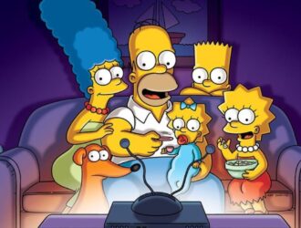A imagem mostra a família Simpson reunida no sofá da sala, assistindo televisão. Homer está ao centro, comendo pipoca, enquanto segura Maggie no colo. Marge, com seus característicos cabelos azuis altos, está sentada ao lado esquerdo dele, abraçando-o. À direita, Lisa aparece animada segurando uma tigela de cereal ou pipoca, e Bart observa curioso por trás de Homer. O cachorro da família, Ajudante de Papai Noel, também está presente, olhando para a tela. Todos estão iluminados pela luz azulada da TV em frente a eles