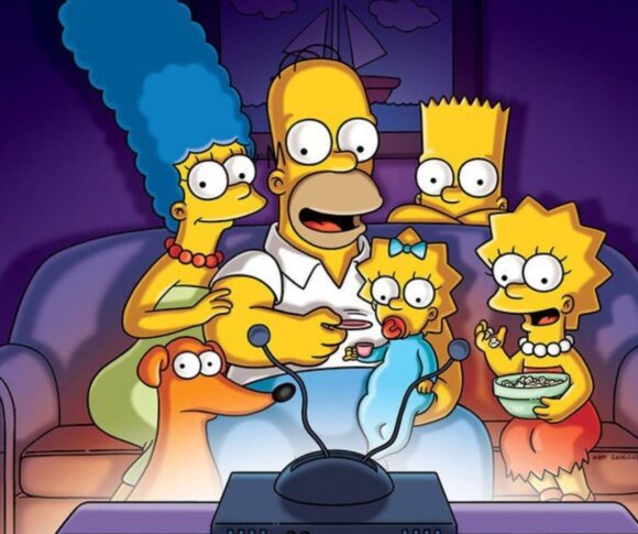 A imagem mostra a família Simpson reunida no sofá da sala, assistindo televisão. Homer está ao centro, comendo pipoca, enquanto segura Maggie no colo. Marge, com seus característicos cabelos azuis altos, está sentada ao lado esquerdo dele, abraçando-o. À direita, Lisa aparece animada segurando uma tigela de cereal ou pipoca, e Bart observa curioso por trás de Homer. O cachorro da família, Ajudante de Papai Noel, também está presente, olhando para a tela. Todos estão iluminados pela luz azulada da TV em frente a eles.