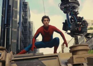 Tom Holland vestido como Homem-Aranha agachado sobre um veículo militar durante as filmagens de uma cena de ação, com um equipamento de câmera suspenso ao lado capturando a cena. Ao fundo, é possível ver prédios modernos e o céu parcialmente nublado.