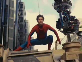 Tom Holland vestido como Homem-Aranha agachado sobre um veículo militar durante as filmagens de uma cena de ação, com um equipamento de câmera suspenso ao lado capturando a cena. Ao fundo, é possível ver prédios modernos e o céu parcialmente nublado
