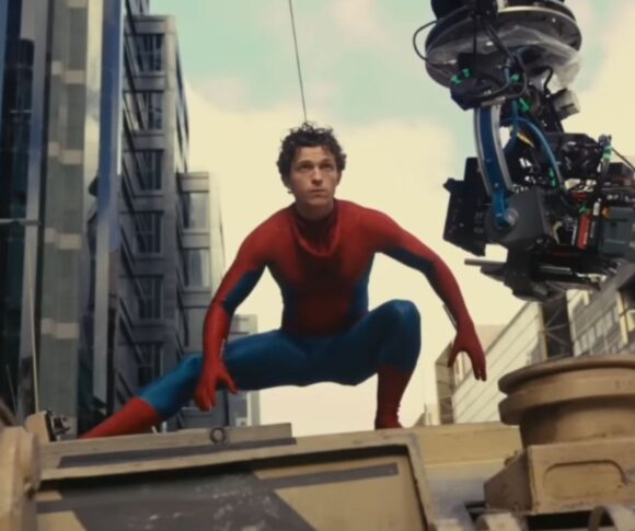 Tom Holland vestido como Homem-Aranha agachado sobre um veículo militar durante as filmagens de uma cena de ação, com um equipamento de câmera suspenso ao lado capturando a cena. Ao fundo, é possível ver prédios modernos e o céu parcialmente nublado.
