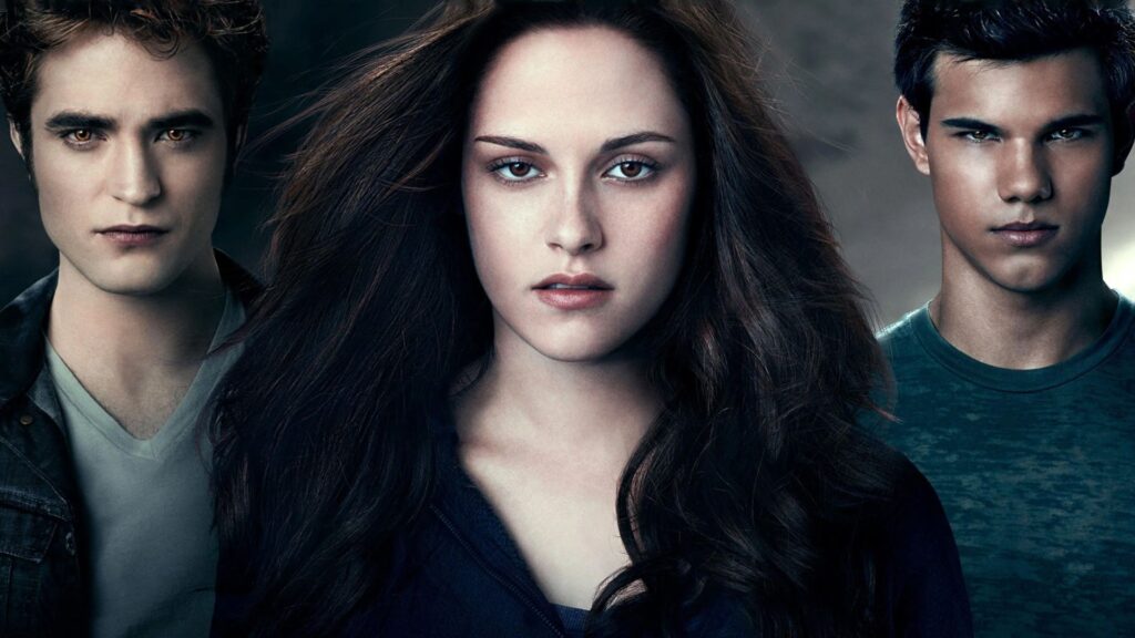Imagem promocional da saga Crepúsculo, com os três personagens principais em destaque. Ao centro está uma jovem de expressão séria, com longos cabelos castanhos soltos, representando Bella Swan. À esquerda, um rapaz de pele muito pálida e olhos dourados, com feições marcantes, representa Edward Cullen. À direita, outro jovem de pele bronzeada e olhar intenso representa Jacob Black. O fundo é escuro e nebuloso, reforçando o clima misterioso e sobrenatural da franquia.