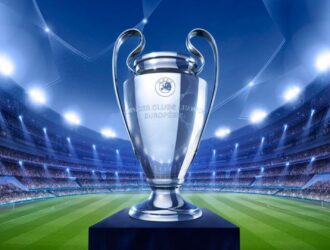 A imagem mostra a taça da Liga dos Campeões da UEFA (Champions League) em destaque no centro. O troféu prateado, conhecido por suas alças grandes e curvas, está apoiado sobre um pedestal azul. Ao fundo, aparece um estádio de futebol iluminado por refletores potentes, com arquibancadas lotadas de torcedores. O campo verde se estende até o horizonte da imagem, criando um clima grandioso e de celebração esportiva