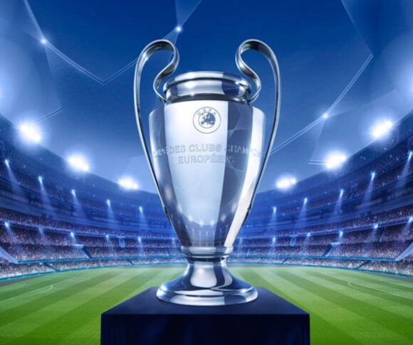 A imagem mostra a taça da Liga dos Campeões da UEFA (Champions League) em destaque no centro. O troféu prateado, conhecido por suas alças grandes e curvas, está apoiado sobre um pedestal azul. Ao fundo, aparece um estádio de futebol iluminado por refletores potentes, com arquibancadas lotadas de torcedores. O campo verde se estende até o horizonte da imagem, criando um clima grandioso e de celebração esportiva