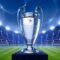 A imagem mostra a taça da Liga dos Campeões da UEFA (Champions League) em destaque no centro. O troféu prateado, conhecido por suas alças grandes e curvas, está apoiado sobre um pedestal azul. Ao fundo, aparece um estádio de futebol iluminado por refletores potentes, com arquibancadas lotadas de torcedores. O campo verde se estende até o horizonte da imagem, criando um clima grandioso e de celebração esportiva