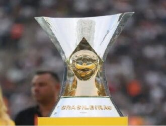 A imagem mostra o troféu do Campeonato Brasileiro Série A, entregue ao time campeão da principal liga de futebol do Brasil. O troféu tem um design moderno e marcante: uma bola dourada no centro, sustentada por uma estrutura metálica com abertura ampla na parte superior. Na base, está gravada a palavra “BRASILEIRÃO”