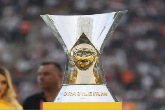 A imagem mostra o troféu do Campeonato Brasileiro Série A, entregue ao time campeão da principal liga de futebol do Brasil. O troféu tem um design moderno e marcante: uma bola dourada no centro, sustentada por uma estrutura metálica com abertura ampla na parte superior. Na base, está gravada a palavra “BRASILEIRÃO”