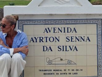Adriane Galisteu está sentada no chão, ao lado de um marco de azulejos que homenageia Ayrton Senna. O monumento exibe a inscrição “Avenida Ayrton Senna da Silva – Tricampeão Mundial de F1 88-90-91 – Residente da Quinta do Lago”, acompanhada de um desenho estilizado de um carro de Fórmula 1. Ela usa calça branca, camisa azul clara com mangas dobradas e óculos escuros. Ao fundo, há um jardim arborizado e construções residenciais, compondo uma cena tranquila e nostálgica.
