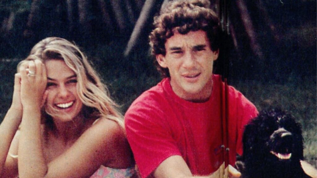 Adriane Galisteu e Ayrton Senna sentados ao ar livre, sorrindo para a câmera. Adriane, à esquerda, está com as mãos juntas apoiando o rosto e cabelo solto, usando biquíni estampado. Ayrton, à direita, veste camiseta vermelha e segura um cachorro preto, que também parece olhar para a câmera.