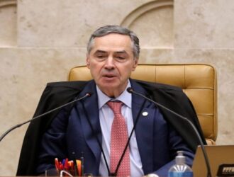 A imagem mostra Luis Roberto Barroso, ex-ministro do Supremo Tribunal Federal (STF) do Brasil, sentado atrás de uma mesa formal. Ele veste uma toga preta judicial sobre um terno com gravata vermelha, e parece estar falando ou se dirigindo a uma audiência, possivelmente durante uma sessão oficial ou julgamento