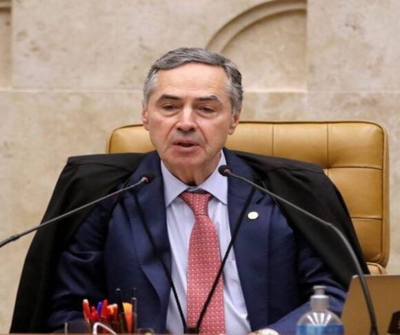A imagem mostra Luis Roberto Barroso, ex-ministro do Supremo Tribunal Federal (STF) do Brasil, sentado atrás de uma mesa formal. Ele veste uma toga preta judicial sobre um terno com gravata vermelha, e parece estar falando ou se dirigindo a uma audiência, possivelmente durante uma sessão oficial ou julgamento