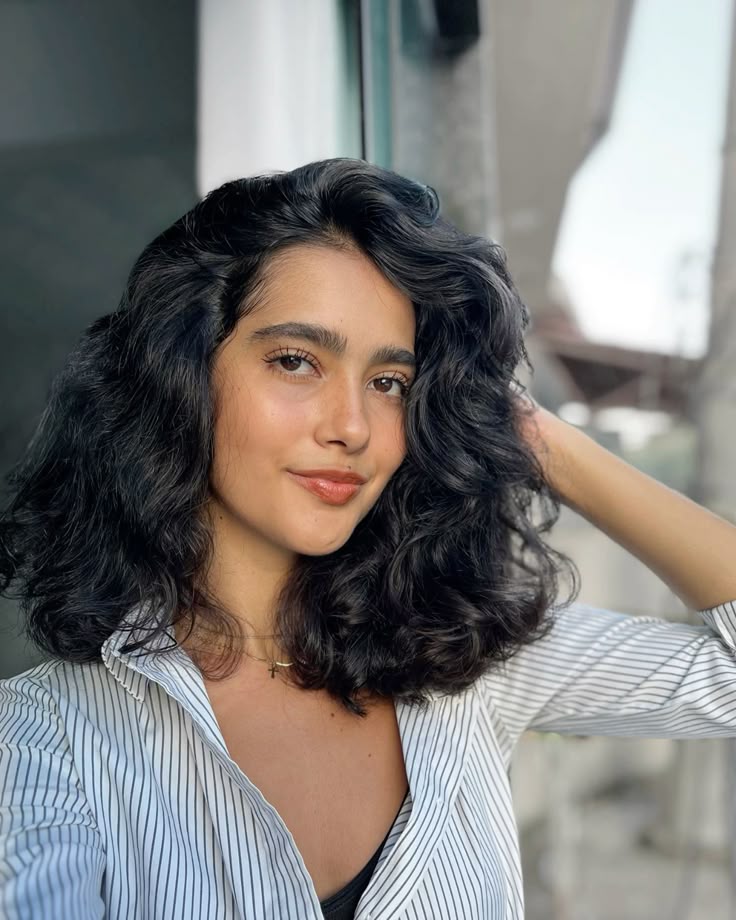 Na imagem aparece uma mulher com cabelos pretos, ondulados, na altura dos ombros e dividido de lado. Ela posa com uma das mãos atrás da cabeça.Ela tem sobrancelhas grossas e maquiagem leve. Usa uma camisa  listrada branca e preto, colares sobrepostos dourados, um deles com pingente de cruz.