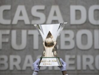 A imagem mostra uma pessoa segurando um grande troféu brilhante, com as duas mãos erguidas, em frente a uma parede com os dizeres “CASA DO FUTEBOL BRASILEIRO”. O troféu tem um globo dourado no centro e está inscrito com “BRASILEIRÃO ASSAÍ”, indicando que é o troféu do Campeonato Brasileiro Série A — a principal liga de futebol do Brasil. O ambiente parece ser a sede da Confederação Brasileira de Futebol (CBF), sugerido pelo texto ao fundo que representa o local como a “Casa do Futebol Brasileiro