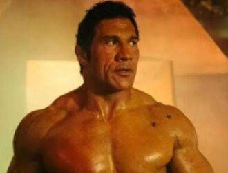 A imagem mostra um Dwayne Johnson, um homem musculoso. Ele interpreta um lutador entrando em um espaço com iluminação dramática e fumaça ao fundo. Veste apenas shorts pretos e luvas brancas, destacando seu físico definido e imponente. O ambiente parece industrial, com tons de laranja e verde na iluminação, criando um clima intenso e cinematográfico. A expressão dele é séria e focada, transmitindo determinação e força