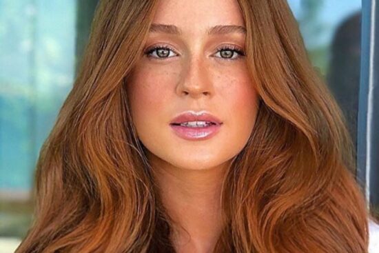 A imagem mostra um retrato da atriz e modelo Marina Ruy Barbosa. Ela tem cabelos ruivos longos e ondulados, e está olhando diretamente para a câmera com um olhar intenso e expressivo. O fundo é desfocado, deixando o foco principal na face de Marina. Ela parece estar usando uma maquiagem natural, com destaque para seus olhos e lábios. A iluminação suave e a composição da imagem criam um efeito elegante e sofisticado, realçando a beleza e o carisma da atriz