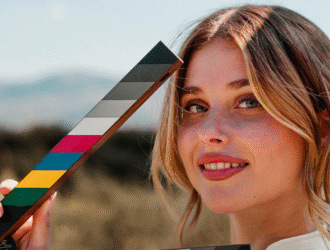 A imagem a atriz espanhola, uma mulher loira e sorridente ao ar livre, segurando um objeto retangular com faixas coloridas e tons de cinza, que lembra um acessório usado em produções audiovisuais para calibrar cores. Ela está em destaque, com fundo desfocado de paisagem natural