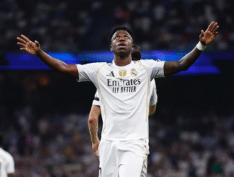 A imagem mostra o jogador de futebol Vinícius Jr., atacante do Real Madrid, comemorando um gol. Ele está com os braços abertos, em uma posição de celebração, vestindo a camisa branca do Real Madrid. O fundo da imagem é um campo de futebol, com a iluminação do estádio