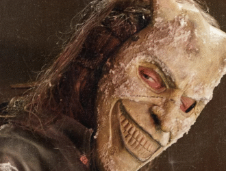 A imagem mostra uma figura macabra usando uma máscara demoníaca com chifres e um sorriso sinistro, parcialmente coberta por neve ou gelo. O olhar vermelho e penetrante reforça o clima de terror e ameaça, remetendo diretamente ao vilão da franquia O Telefone Preto