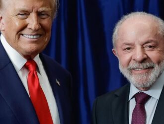 A imagem registra um momento diplomático entre os presidentes Donald Trump, dos Estados Unidos, e Luiz Inácio Lula da Silva, do Brasil. Ambos aparecem em frente a uma cortina azul escura, sorrindo e apertando as mãos em gesto de cordialidade. Trump veste um terno azul com camisa branca e gravata vermelha, enquanto Lula usa um terno escuro com camisa azul clara e gravata vinho.