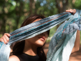 A imagem mostra uma pessoa ao ar livre, em meio a um ambiente natural com árvores ao fundo. Ela segura um lenço azul claro com estampas e detalhes dourados e avermelhados, esticando-o à frente do rosto, parcialmente cobrindo os olhos. A luz natural atravessa o tecido, criando um efeito suave e artístico. A cena transmite uma sensação de leveza, introspecção e conexão com a natureza