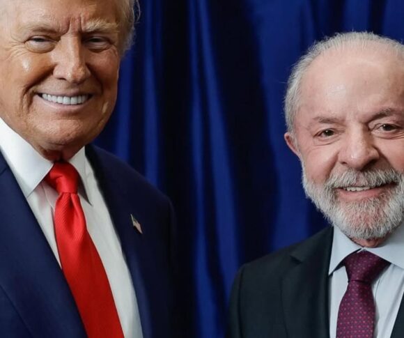 A imagem registra um momento diplomático entre os presidentes Donald Trump, dos Estados Unidos, e Luiz Inácio Lula da Silva, do Brasil. Ambos aparecem em frente a uma cortina azul escura, sorrindo e apertando as mãos em gesto de cordialidade. Trump veste um terno azul com camisa branca e gravata vermelha, enquanto Lula usa um terno escuro com camisa azul clara e gravata vinho.
