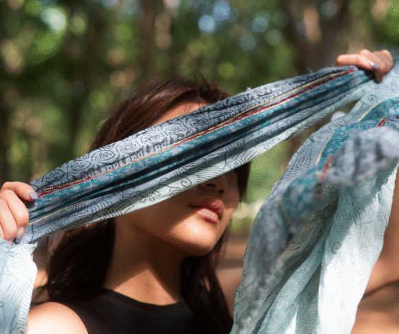 A imagem mostra uma pessoa ao ar livre, em meio a um ambiente natural com árvores ao fundo. Ela segura um lenço azul claro com estampas e detalhes dourados e avermelhados, esticando-o à frente do rosto, parcialmente cobrindo os olhos. A luz natural atravessa o tecido, criando um efeito suave e artístico. A cena transmite uma sensação de leveza, introspecção e conexão com a natureza