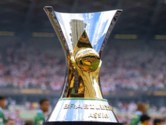 A 12 rodadas do fim A imagem mostra o troféu da Copa do Mundo da FIFA, um dos maiores símbolos do futebol mundial. O troféu é feito de ouro maciço e possui uma forma característica, com duas figuras humanas segurando o globo terrestre. Ele está sendo exibido em um estádio de futebol, provavelmente durante uma partida ou cerimônia da Copa do Mundo. O fundo escuro sugere que a imagem foi capturada durante a noite ou em um ambiente com pouca iluminação, destacando ainda mais o brilho e a imponência do troféu