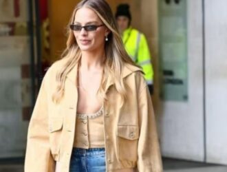 A imagem mostra uma mulher loira de cabelos lisos caminhando em ambiente urbano. Ela usa óculos de sol pequenos, brincos de pérola, top bege com detalhe brilhante, jaqueta cropped bege clara e calça jeans azul. O look transmite um estilo moderno e casual-chic. Ao fundo, há uma pessoa de colete amarelo fluorescente e parte de um prédio