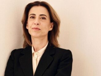 A imagem mostra uma mulher em pé, de frente, com expressão séria e postura confiante. Ela tem cabelos castanhos claros, lisos e na altura dos ombros. Veste um blazer preto sobre uma camisa clara, abotoada até o pescoço, transmitindo um ar sofisticado e profissional. O fundo é liso e claro, o que destaca a figura da mulher e reforça o tom sóbrio e elegante da fotografia