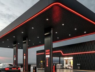 A imagem mostra um posto de combustível moderno, com design futurista e iluminação em tons de vermelho e branco. A estrutura principal é preta, com acabamento metálico e linhas geométricas. O teto da cobertura tem luzes embutidas e uma borda iluminada em vermelho que contorna todo o espaço. A loja de conveniência, ao fundo, segue o mesmo estilo arquitetônico, com paredes pretas texturizadas e faixas de luz vermelha. Um carro esportivo preto está estacionado próximo às bombas de combustível. O céu está nublado, sugerindo o fim de tarde ou início da noite, o que realça o contraste das luzes com o ambiente