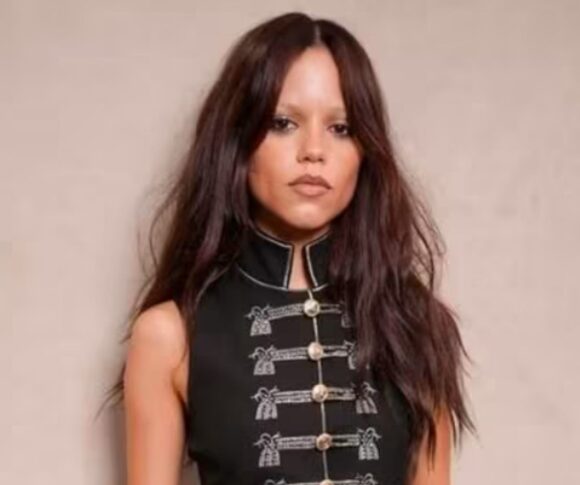 Jenna Ortega (mulher, pele clara, cabelos castanhos longos) aparece de pé vestindo mini saia jeans, blusa preta com botões dourados, escarpins pretos