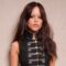 Jenna Ortega (mulher, pele clara, cabelos castanhos longos) aparece de pé vestindo mini saia jeans, blusa preta com botões dourados, escarpins pretos