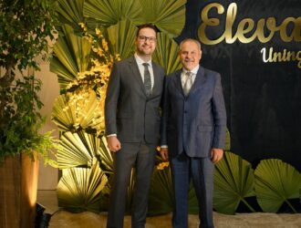A imagem mostra dois homens posando lado a lado em um cenário elegante de evento. Ambos vestem ternos formais: o homem à esquerda usa terno cinza com camisa branca e gravata listrada, enquanto o homem à direita usa terno azul com camisa branca e gravata clara. Ao fundo, há uma parede escura com o logotipo dourado “Eleva Uningá”, e uma decoração composta por grandes folhas verdes e arranjos de flores amarelas iluminados por luz quente. À esquerda, há um grande vaso de madeira com plantas verdes, contribuindo para o ambiente sofisticado e natural da cena