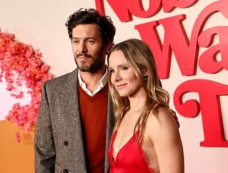A imagem mostra um casal posando juntos em um tapete vermelho, diante de um painel decorado com flores cor-de-rosa e letras grandes em vermelho. O homem veste um casaco cinza com padronagem discreta sobre um suéter vermelho e camisa branca. Ele tem barba curta e cabelo castanho escuro levemente ondulado. A mulher ao lado usa um vestido vermelho com decote profundo e alças finas, de tecido acetinado. Ela tem cabelo loiro, solto e levemente ondulado, e usa maquiagem suave em tons naturais. Ambos sorriem levemente e olham na direção das câmeras, em um ambiente iluminado e elegante típico de eventos de estreia ou premiação