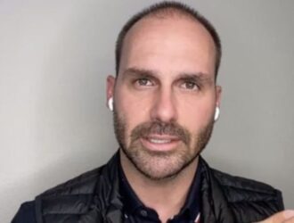 A imagem mostra um homem em plano médio, posicionado contra um fundo neutro de cor clara. Ele tem barba curta, cabelos ralos na parte superior da cabeça e está usando fones de ouvido brancos sem fio. Veste uma camisa escura e um colete preto acolchoado. Sua expressão facial é séria, e ele parece estar falando ou explicando algo, possivelmente em uma transmissão ao vivo ou gravação de vídeo