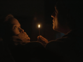 A imagem mostra uma cena sombria e tensa, iluminada apenas pela chama de um fósforo. À esquerda, uma mulher assustada tem a boca tapada por uma mão, sugerindo que está sendo contida ou silenciada. À direita, um homem observa-a de perto enquanto segura o fósforo aceso. A iluminação dramática e o clima de suspense indicam que se trata de um momento de perigo ou ameaça